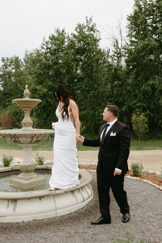 Bride with long curly brown hair walking on a foundation ledge in her white dress, holding her groom’s hand, with lush green grass in the background